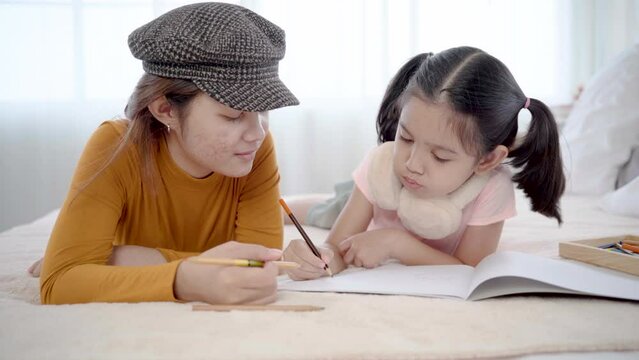 Asian elder girl spent holiday with cute girl lying on bed painting together in holiday. adorable girl drawing portrait imagination in paper, young girl teaching preschool kid sibling relationship
