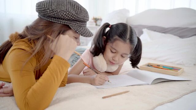 Elder sister and cute little sister lying on bed in bedroom painting together in holiday. adorable girl drawing her sister in paper, portrait painting, child teaching concept.
