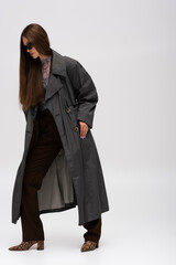 full length of teenage model in sunglasses and trench coat posing on gray.