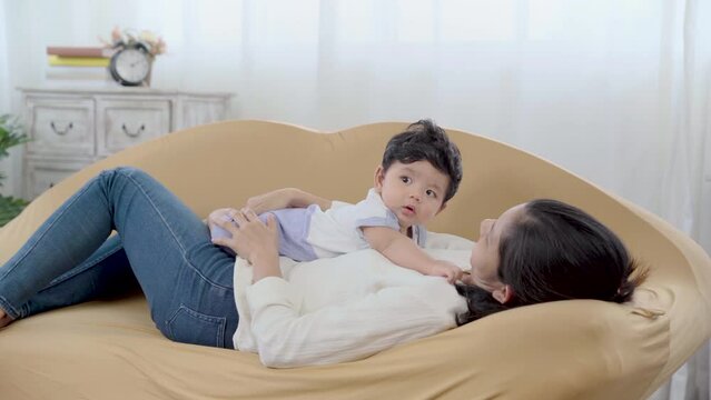 Healthy asian mother play and tease with newborn baby in living room, mom holding lifting toddler on brown sofa with fun and tender. Adorable little boy having fun with mum togerther at home.
