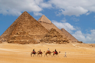 Cairo, Egypt. 08.25.2022. Group of tourists riding camels seeing the pyramids of Menkaure, Chephren and Cheops. - obrazy, fototapety, plakaty