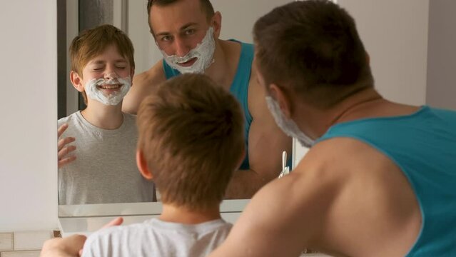 Father and son with shaving foam on face smiling and fool around in front of reflection in bathroom mirror.Time together