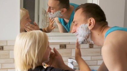 Father and son fool around in bathroom. Little boy laughs and smears shaving foam on dads face