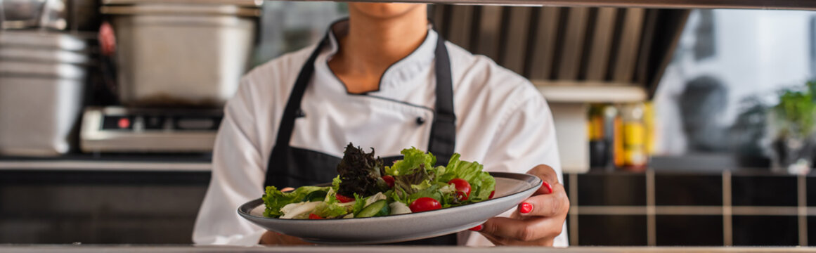 cropped view of african american chef holding plate with freshly cooked salad in professional kitchen, banner.