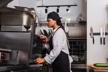 side view of young african american chef in hat using convection oven in kitchen.