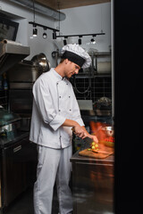 young chef in uniform cutting fresh cucumber behind glass door in professional kitchen.