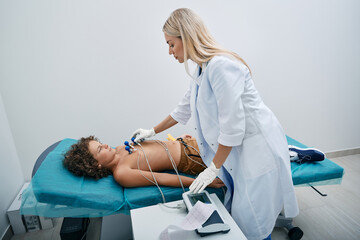Cardiologist installing vacuum sensors on male child chest during electrocardiogram procedure at medical clinic. ECG for children