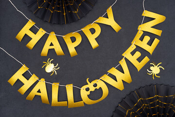 Happy Halloween card. Golden words as a garland on a dark background decorated with black paper fans, gold cobweb and shiny spiders. The Scary Halloween background.