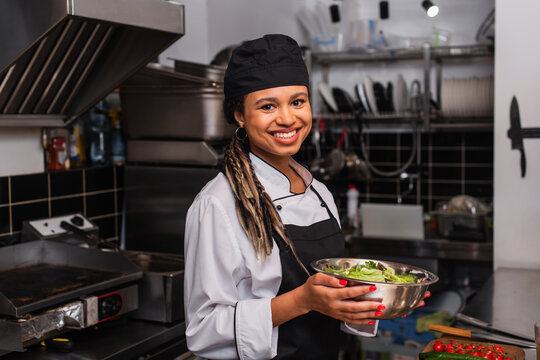 happy and young african american chef holding bowl with salad in professional kitchen.
