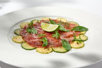 Beef carpaccio with zucchini and cucumber on white table with sunlight shadows. Appetizer of meat - beef carpaccio on summer dining. Sliced raw meat in italian style. Aesthetic menu.