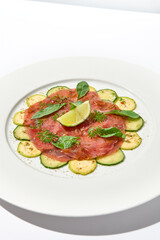 Beef carpaccio with zucchini and cucumber on white table with sunlight shadows. Appetizer of meat - beef carpaccio on summer dining. Sliced raw meat in italian style. Aesthetic menu.