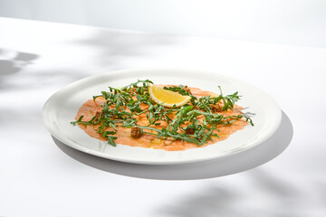 Salmon carpaccio with arugula on white background in summer day. Fish appetizer - salmon carpaccio in fine dining with shadows. Elegant carpaccio of raw salmon and sauce. Italian food. Aesthetic menu.