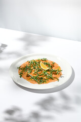 Salmon carpaccio with arugula on white background in summer day. Fish appetizer - salmon carpaccio in fine dining with shadows. Elegant carpaccio of raw salmon and sauce. Italian food. Aesthetic menu.