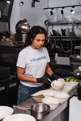 tattooed african american volunteer in t-shirt with lettering holding plastic container with soup and fork in kitchen.