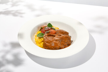 Traditional italian dish - osso buco with risotto milanese. Stew meat on bone with rice on white plate. Summer italian dining. Italian osso buco with garnish with hard shadows