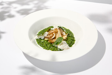 Green risotto with squid and stracciatella cheese on white plate. Creamy risotto with spinach, calamari and creamy cheese. Italian risotto with squid on light background with shadows of leaves