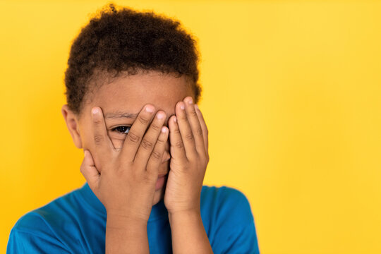 Close-up of terrified preteen boy covering face with hands. Mixed race child wearing blue T-shirt looking at camera in fear against yellow background. Home violence and children phobia concept