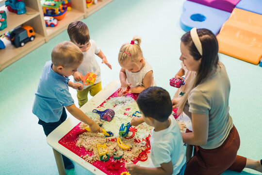 Calming sensory play with moldable kinetic sand at nursery school. Toddlers with their teacher having fun around the table using different tools for sculpting sand such as colorful and textured