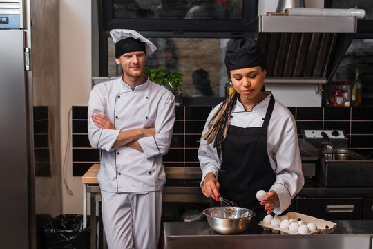 young african american sous chef in apron holding egg near bowl and whisk while cooking near chef in kitchen. - Powered by Adobe