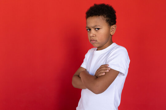 Portrait of resentful preteen boy wearing white T-shirt standing with folded arms. Mixed race child wearing white T-shirt looking at camera against red background. Resentment concept