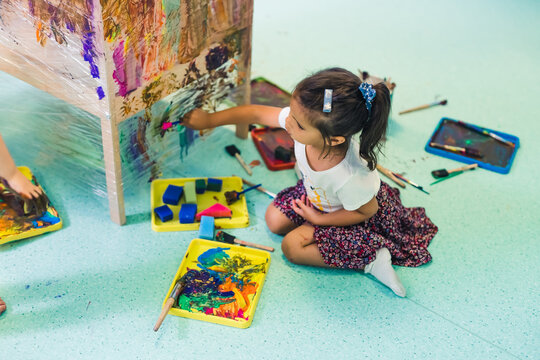 Cling film painting. Little girl toddler painting with a sponge and paints on a cling film wrapped all the way round the wooden shelf unit. Creative activity for kids development at the nursery school