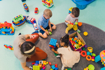 Toddlers and their nursery teacher playing with plastic building blocks and colorful car toys while sitting on the floor in a playroom. Early brain and skills development. High quality photo - obrazy, fototapety, plakaty