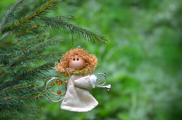 Fototapeta premium Angel with trombone on a Christmas tree branch. Christmas tree decoration angel-girl in beige dress hangs on pine tree branch. Free copy space. Christmas, Angel day or New Year greeting .