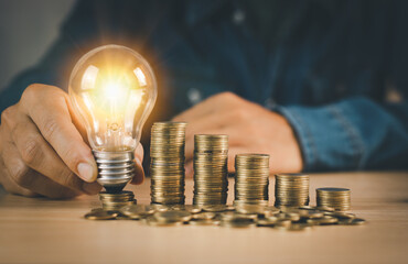 Man's hand holding light bulb with stacked golden coins on wooden table, energy saving and money...