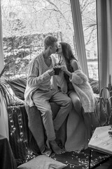 A young and attractive couple, a guy and a girl, are sitting on the windowsill in blankets and warm sweaters in winter during a snowfall, hugging and drinking tea. Emotions and feelings