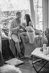 A young and attractive couple, a guy and a girl, are sitting on the windowsill in blankets and warm sweaters in winter during a snowfall, hugging and drinking tea. Emotions and feelings