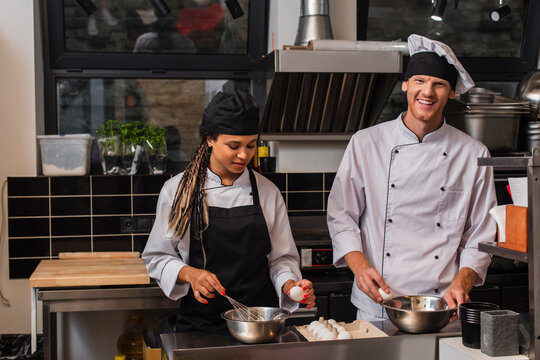 young african american sous chef in apron holding egg near bowl and whisk while cooking near happy chef in kitchen.