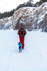 Fototapeta na wymiar Rear view young man in red brown warm clothes with backpack with mixed breed dog in warm blue suit walking in snow among rocks and cliffs in winter Active lifestyle hiking pets adoption