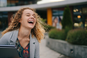 Funny happy caucasian young woman with curly hair wearing jacket laughing standing near city street building. School education. Attractive beautiful girl. Youth