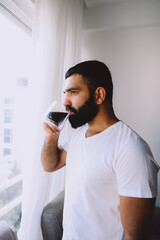 Stylish bearded male drinking black coffee out of transparent cup wearing white shirt at home standing by window