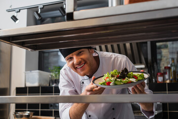 cheerful chef holding plate with freshly cooked salad in professional kitchen.