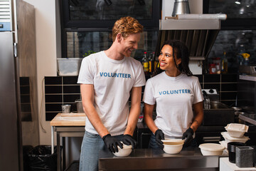 Obraz premium happy multiethnic volunteers in t-shirts preparing plastic containers with food in kitchen.