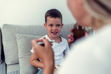 Schoolboy sick and home, willing to take some syrup from mom. Little boy taking a spoon of medicine.
