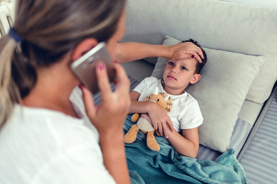 Photo Of Worried Mother Checking Temperature Of Her Little Son Who Is Lying In Bed With Fever. Young Mother Checking The Temperature Of Her Ill 6 Years Old Boy On Couch, Calling Doctor.