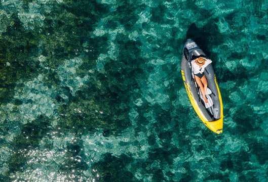 A lonely female in a straw hat relaxing lying in floating inflatable kayak on the turquoise Adriatic Sea waves. Aerial coastal top view shot. Exotic countries vacations concept.
