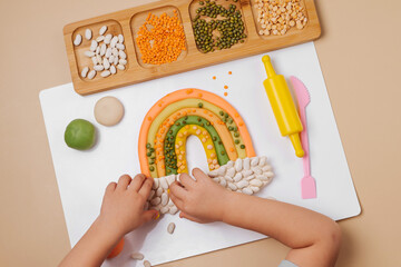 Child hands creating rainbow from play dough for modeling with decorate from dried beans. Art...