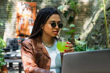 young african american freelancer in sunglasses using laptop and holding glass with cocktail in outdoor terrace. - obrazy, fototapety, plakaty