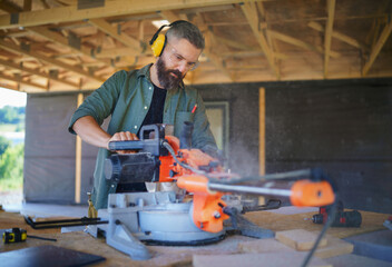 Construction worker working with eletric saw inside wooden construction of house, diy eco-friendly homes concept.