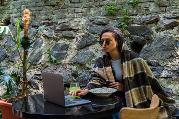 Fototapeta premium african american freelancer in sunglasses covered in blanket using laptop and having lunch in cafe on terrace.