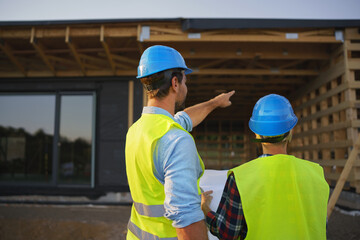 Construction engineers or architects with blueprints checking eco building site of wood frame house