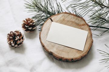 Christmas stationery still life. Blank business card, invitation mockup on cut wooden round board. Pine cones and green Christmas tree branches on white linen cloth background. Top view, no people.
