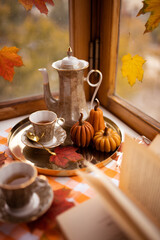 Autumn still life with candles in the shape of pumpkins, tea cup, autumn leaves on the window, teapot, golden tray and vintage book