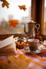 Autumn still life with candles in the shape of pumpkins, tea cup, autumn leaves on the window, teapot, golden tray and vintage book