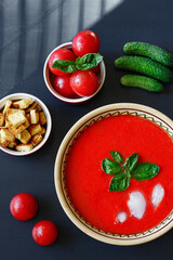 Red Spanish vegetable cold cream soup gazpacho in plate with basil leaves, ice cubes in it, ingredients around: tomatoes, cucumbers, croutons; shadow from blinds, flatlay, vertical, dark background