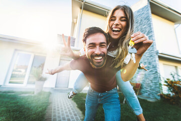 Happy young couple holding home keys after buying real estate - Husband and wife standing outside in front of their new house - 530086905