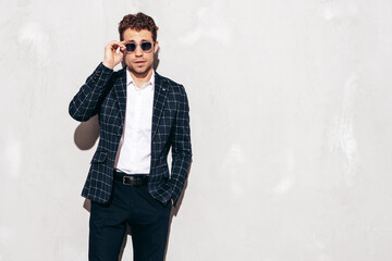 Portrait of handsome confident stylish hipster lambersexual model. Sexy modern man dressed in black elegant suit. Fashion male posing in studio near grey wall. In sunglasses